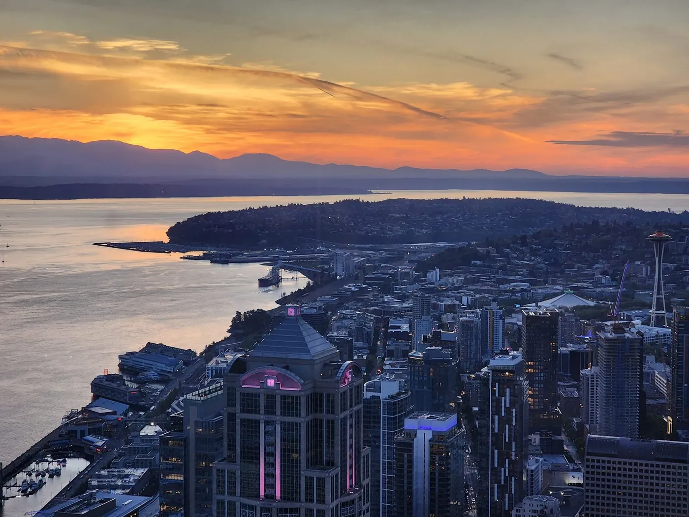 An aerial view of Seattle at summer sunset — Elliott Bay and the Olympic Mountains under a burning orange sky, the Space Needle at right